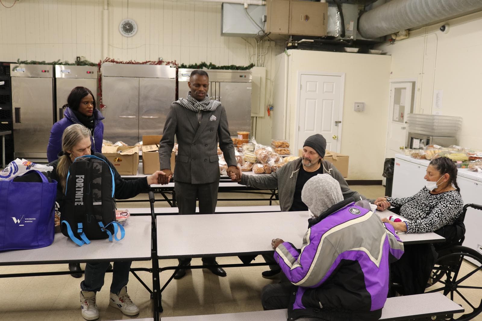 Pastor Henry Edward Ssuuna holding hands in prayer with guests seated around tables at a community meal